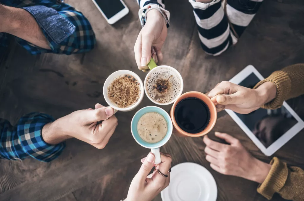 Petit-déjeuner d'onboarding. Vue du dessus de plusieurs personnes réunies autour d’une table, levant des tasses de café de couleurs différentes pour un moment convivial en entreprise, avec smartphones et tablette posés sur la table.