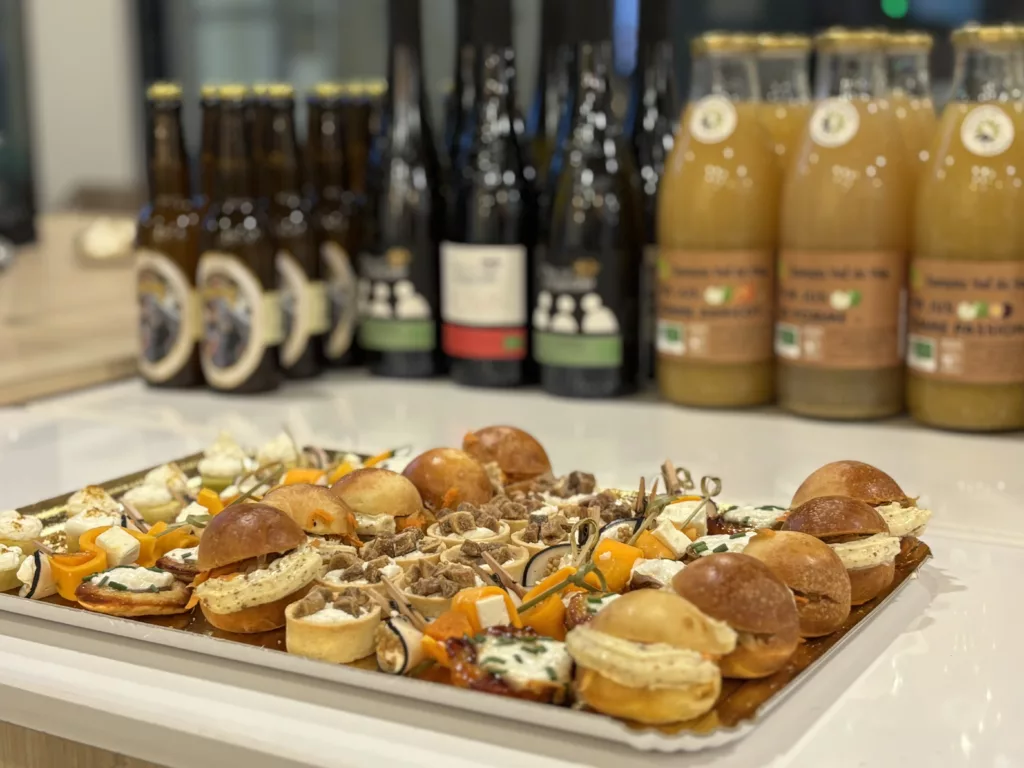 Plateau de bouchées apéritives salées composé de mini-burgers, tartelettes, fromages et légumes rôtis, posé sur un comptoir avec des bouteilles de boissons en arrière-plan. Réduire l’empreinte carbone de vos événements
